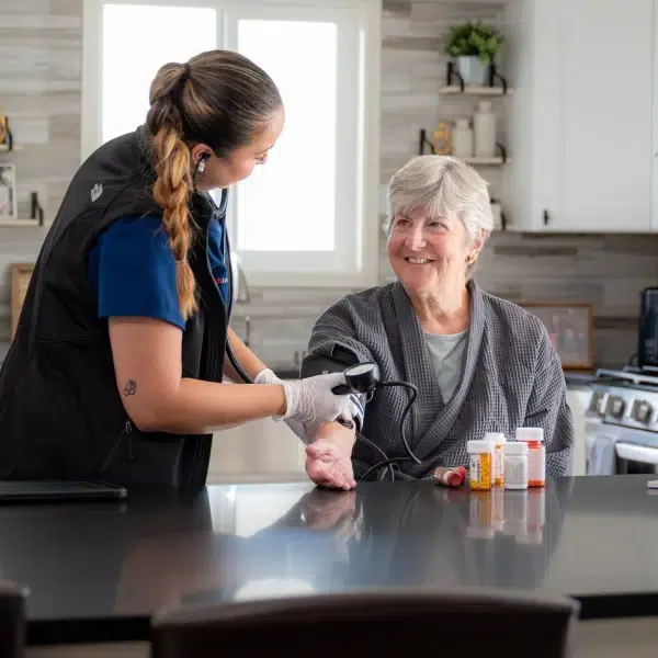 DispatchHealth provider in blue uniform checking vital signs of a smiling senior woman at her kitchen counter, with prescription bottles visible nearby