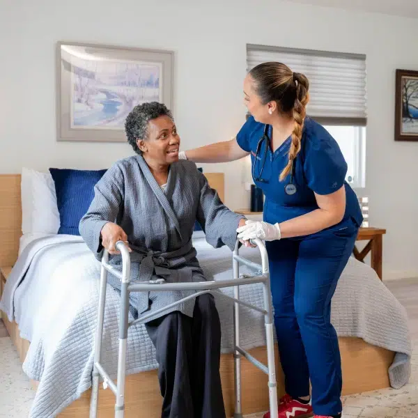 DispatchHealth nurse in blue scrubs providing compassionate care to elderly patient using walker in bedroom during home visit
