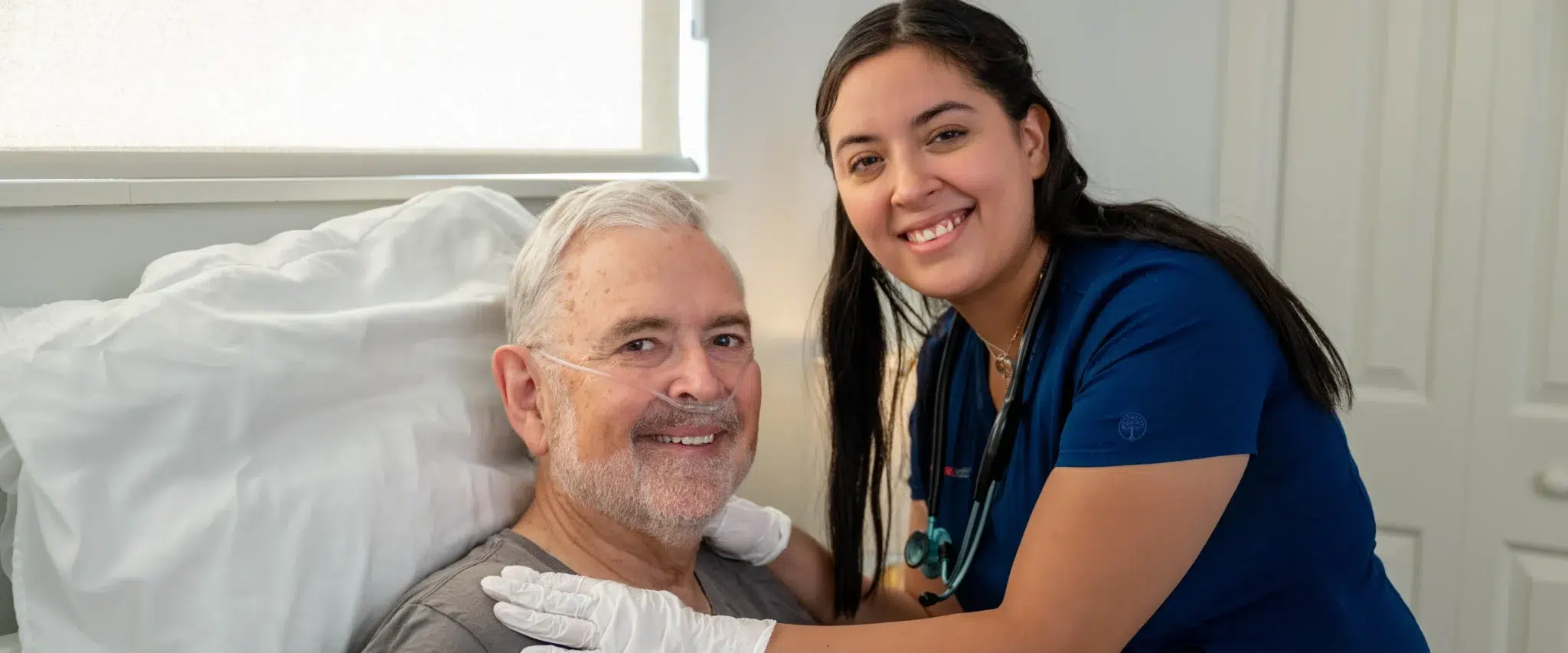 Smiling DispatchHealth provider in blue scrubs with stethoscope caring for elderly male patient with oxygen support in hospital bed