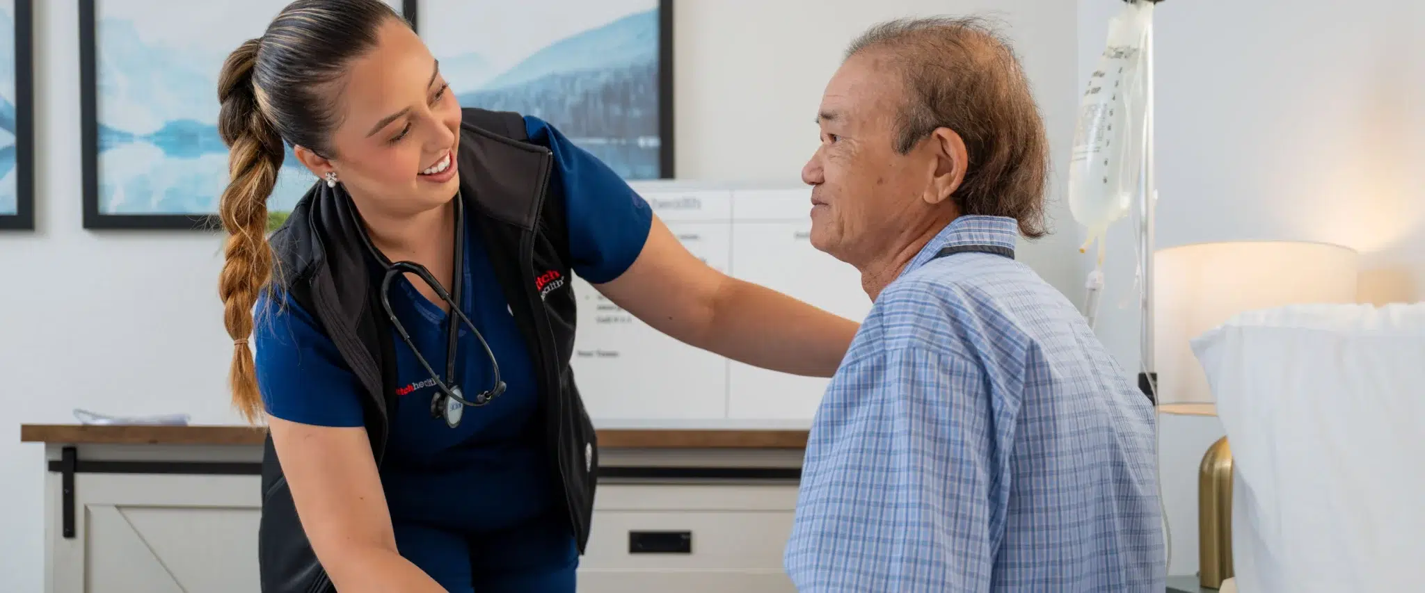 DispatchHealth provider in blue scrubs examining elderly male patient in medical office setting with caring touch on shoulder