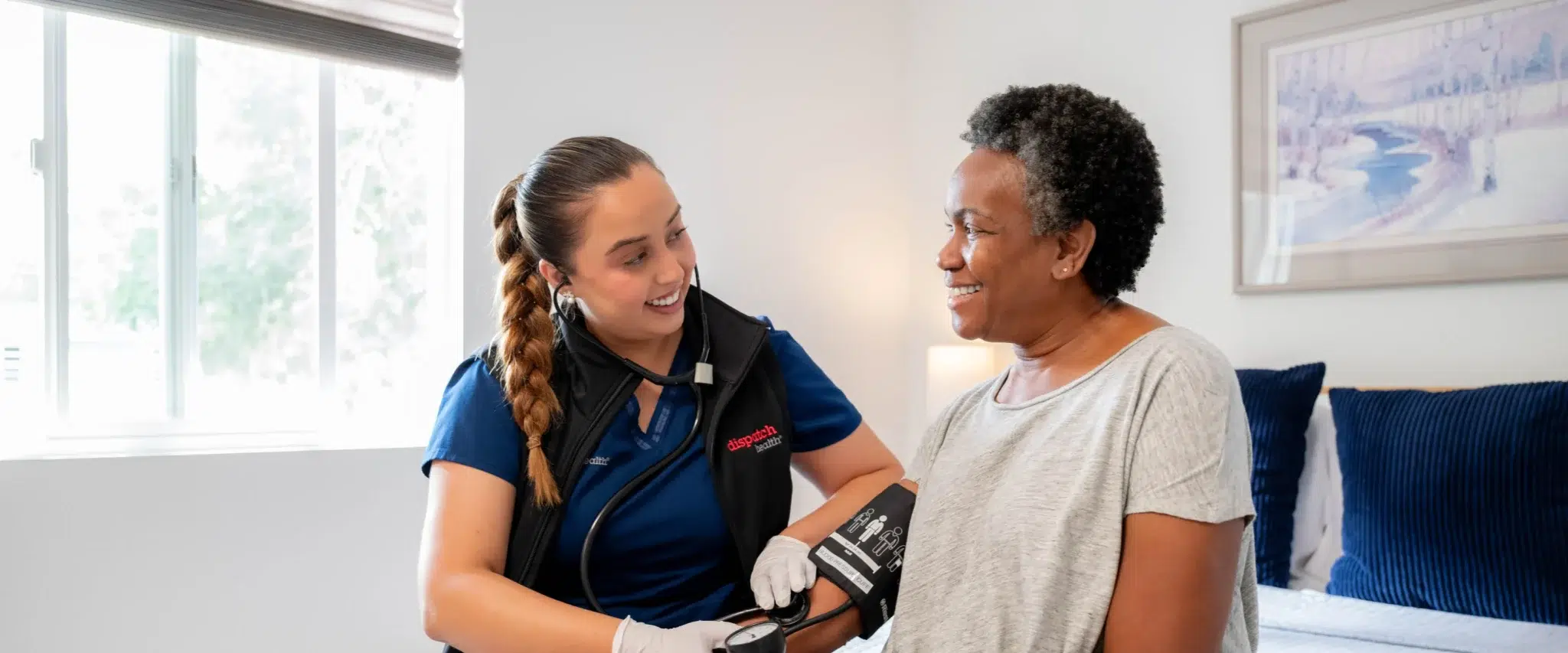 DispatchHealth provider in blue scrubs checking blood pressure of elderly African American female patient in bright modern bedroom