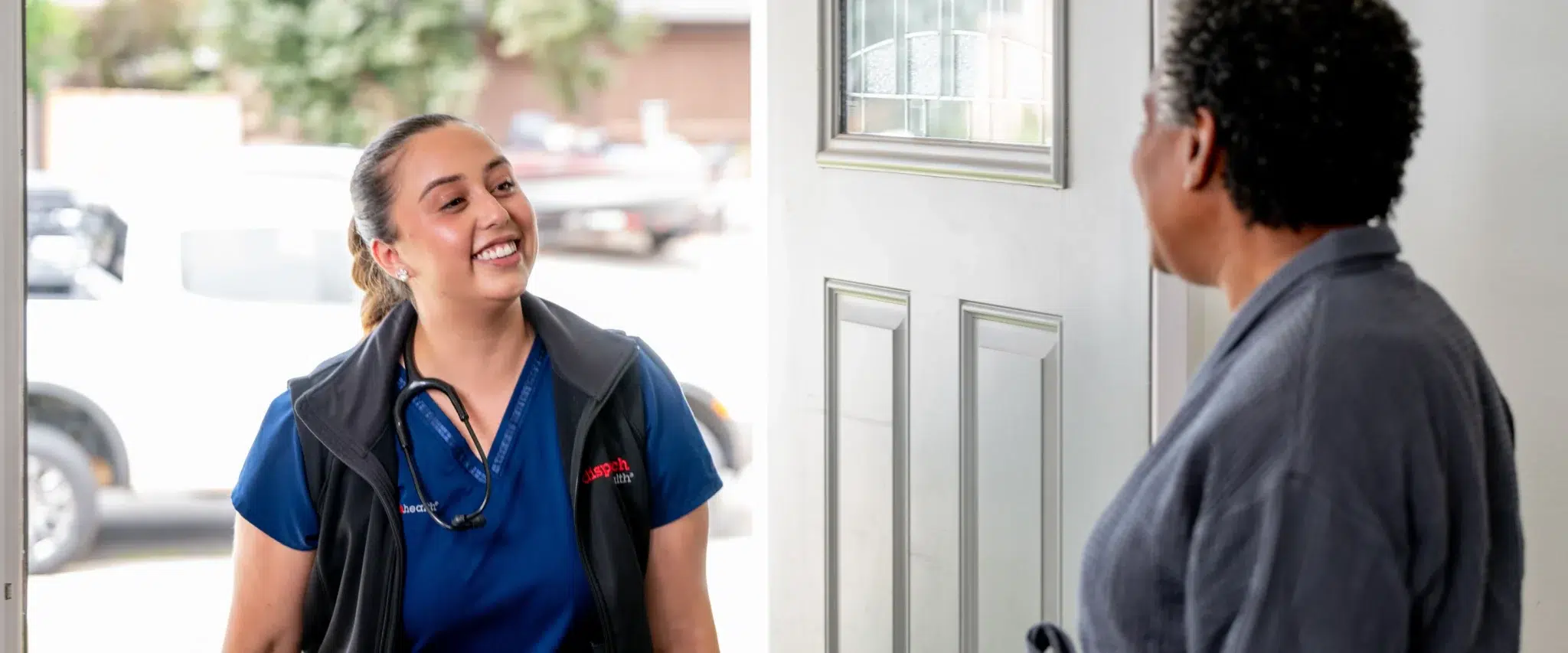Friendly DispatchHealth provider in blue scrubs with stethoscope smiling at doorway greeting patient