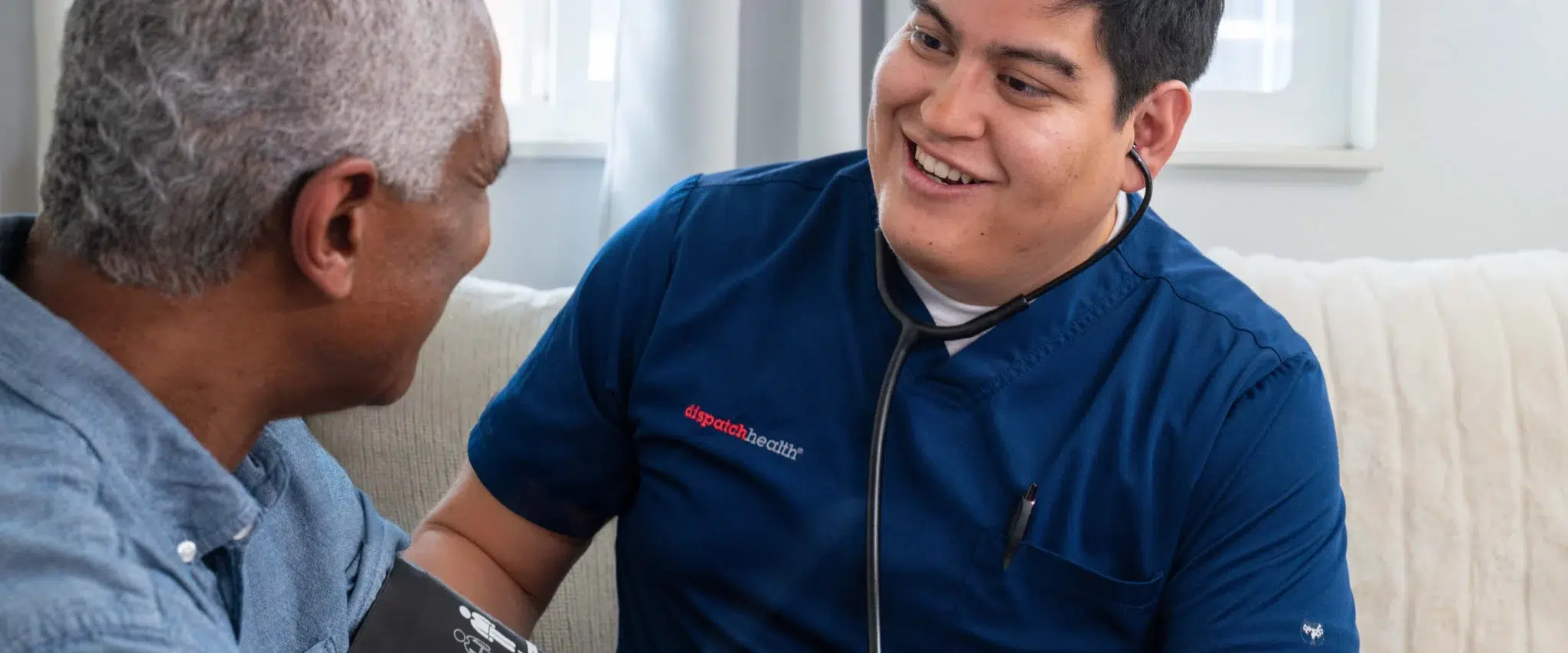 Cheerful DispatchHealth provider in blue scrubs with stethoscope sharing a joyful moment with elderly male patient during home visit