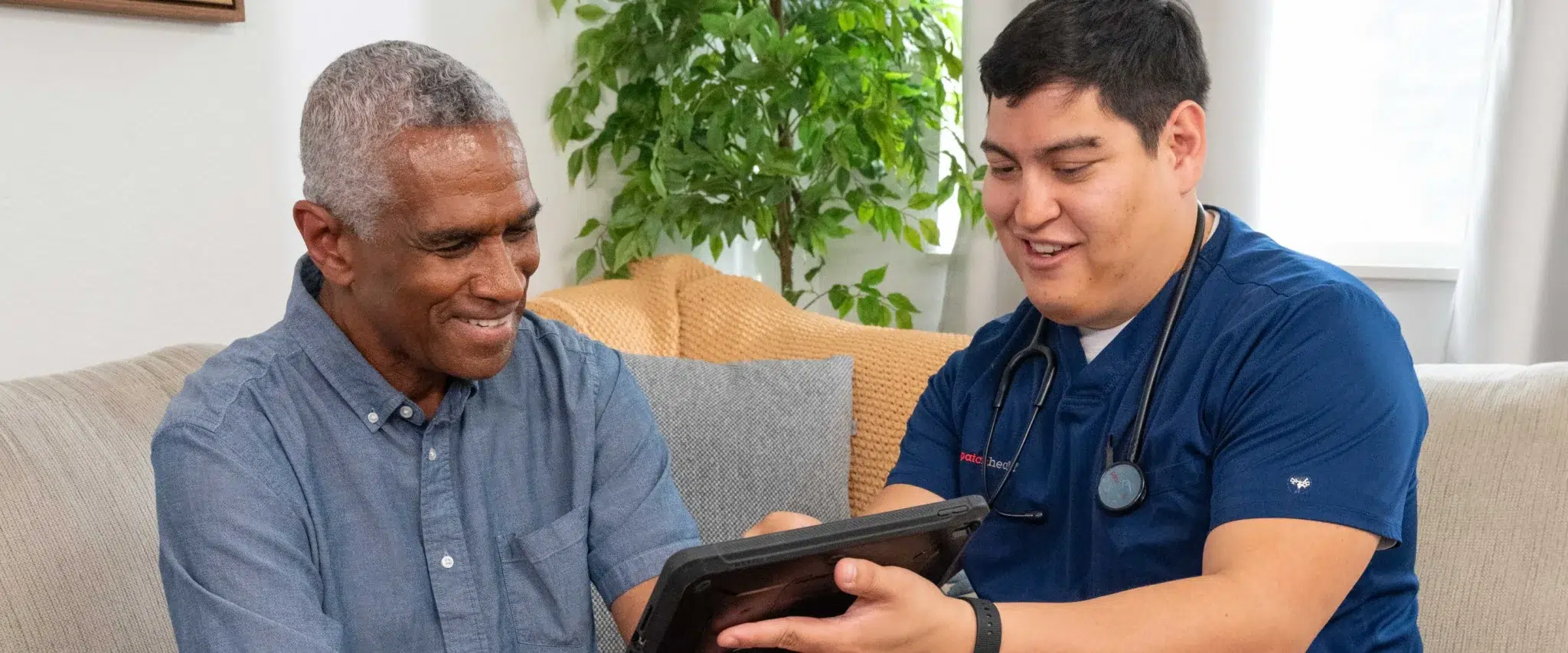DispatchHealth provider in blue scrubs showing tablet to smiling elderly male patient on living room couch during home healthcare visit