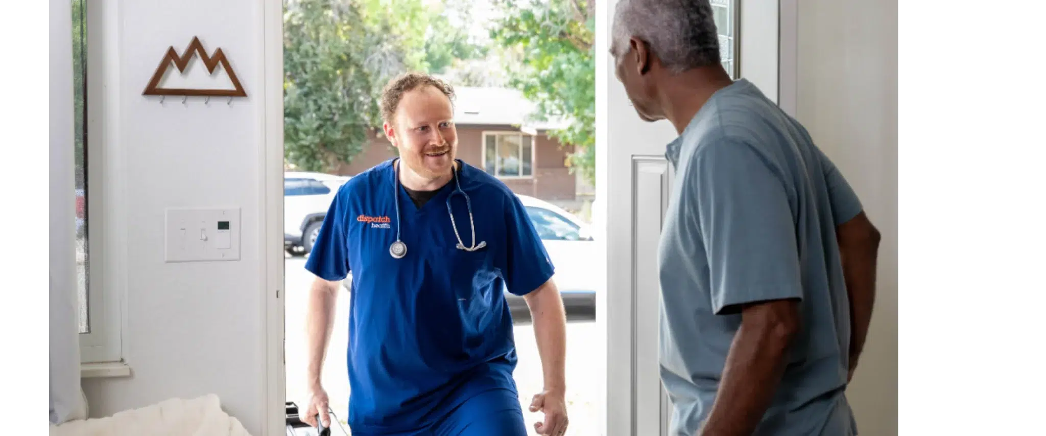 DispatchHealth provider in blue scrubs with stethoscope greeting elderly male patient at front door of home with friendly smile