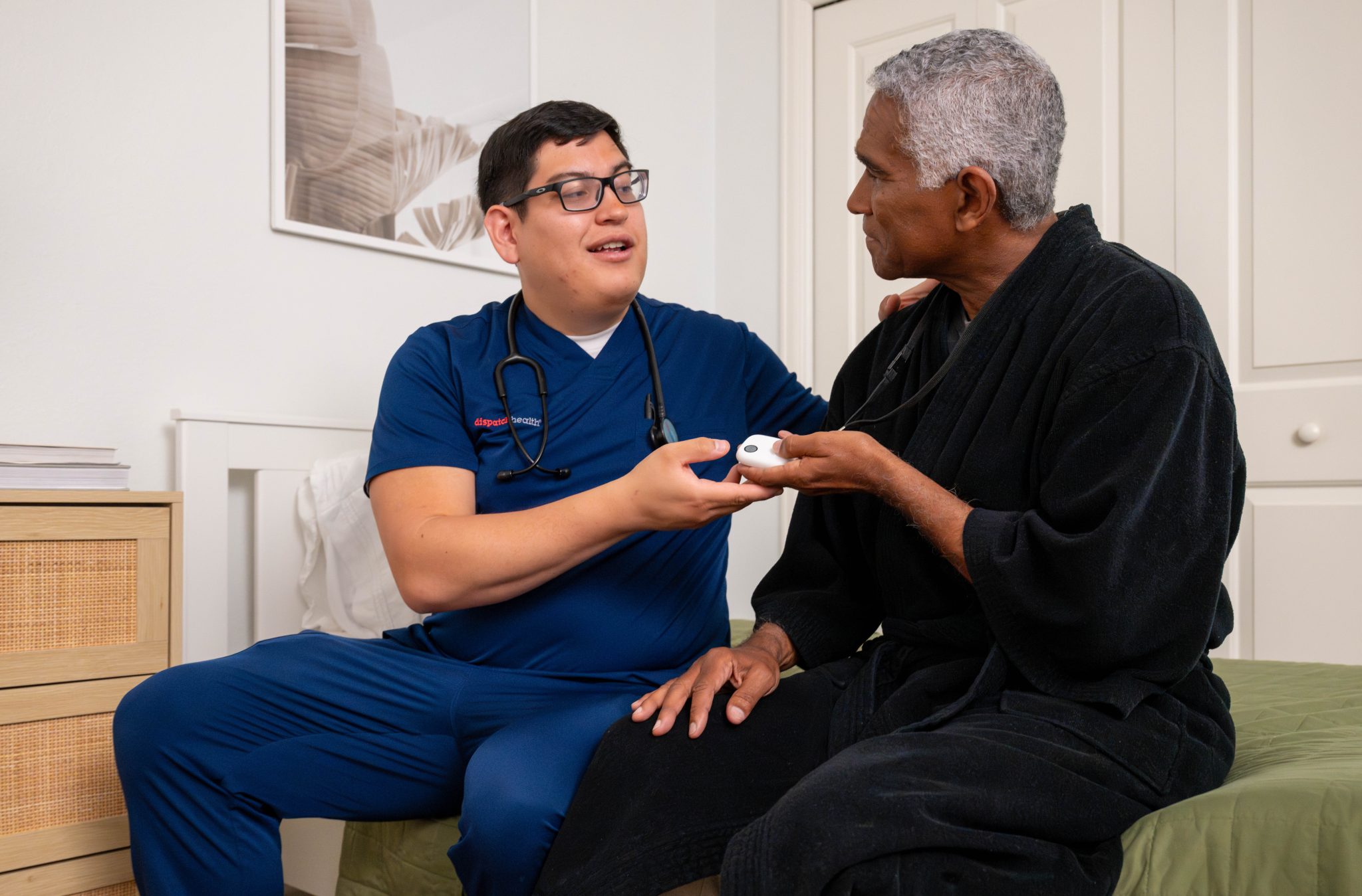 DispatchHealth healthcare provider in blue scrubs sitting with an elderly patient in a bedroom, showing him a medical device while engaged in friendly conversation