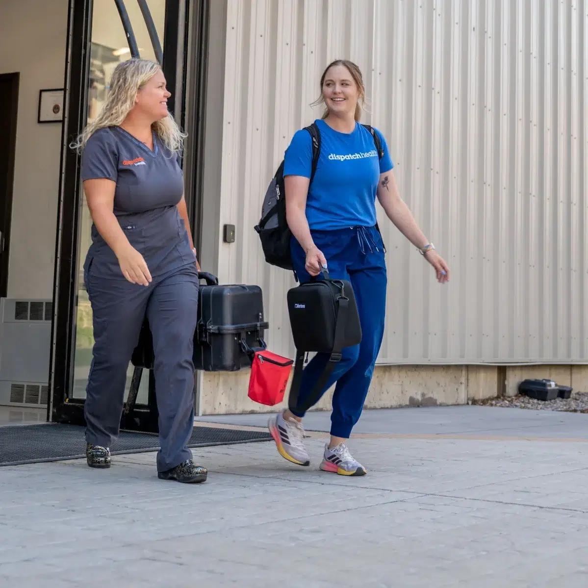 Two DispatchHealth medical professionals carrying equipment bags exiting modern medical facility for mobile healthcare visit